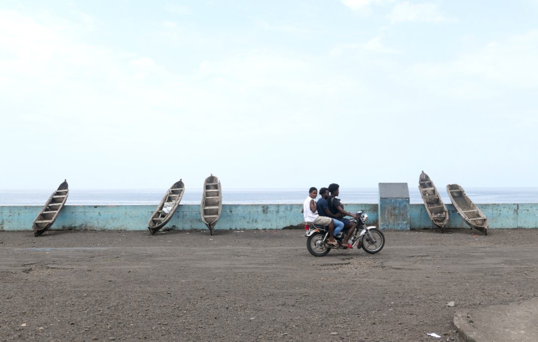 Drei Männer auf einem Moped, dahinter das Meer